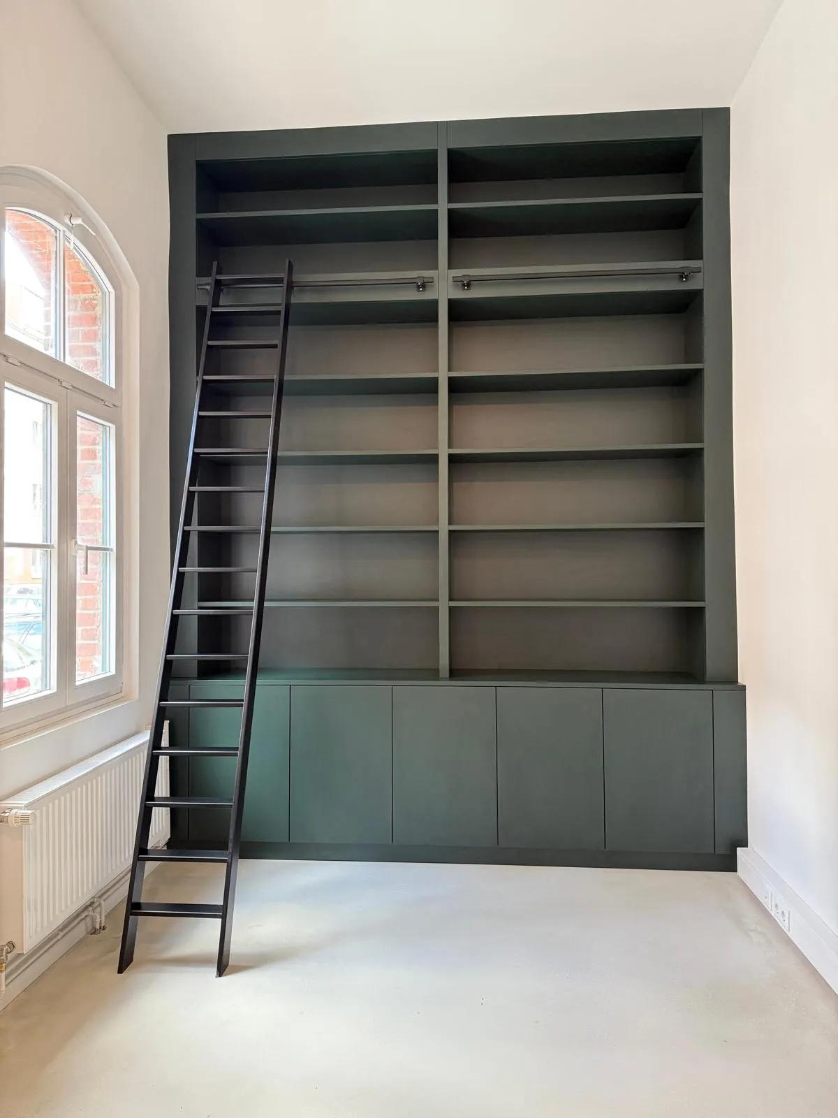 Eine dunkelgrüne Regalwand mit Leiter bietet offene Fächer für eine große Büchersammlung.